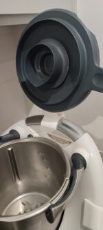 Pokrywa Thermomix TM6 uniesiona nad misą, oparta o uchwyt założony na łuk z tyłu urządzenia – widok z boku