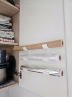 Trzy rolki – papier do pieczenia, folia spożywcza i folia aluminiowa – w kołyskach dwóch elementów na drzwiczkach szafki
